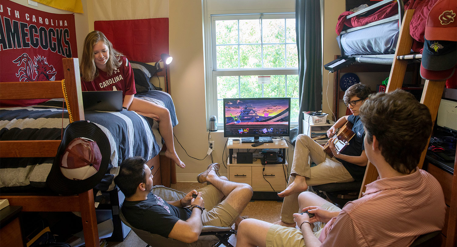 Four students hanging out in a residence hall playing a video game and guitar.