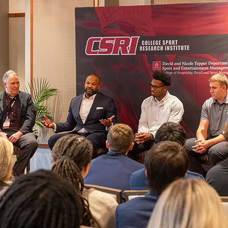 Four panelists participate in a discussion at a College Sport Research Institute event as audience members listen.