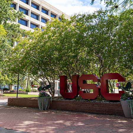 Large garnet USC letters stand outside a University of South Carolina campus building, surrounded by trees and landscaping.