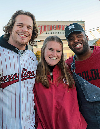 Three alumni wearing Carolina gear at a tailgate with Williams-Brice Stadium behind them. 